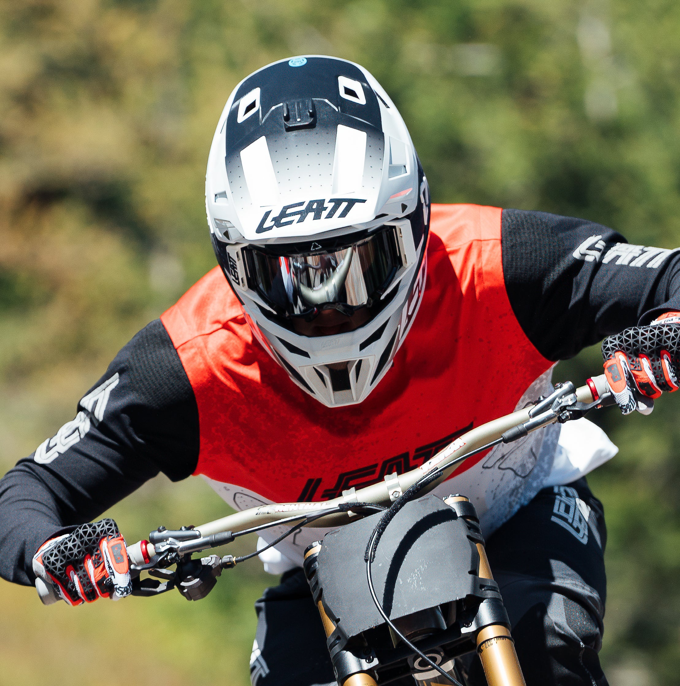 A mountain biker wearing a Leatt full-face helmet, black and red Leatt jersey, and gloves riding a dirt bike on a blurred outdoor trail.